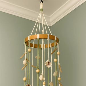 以下が含まれることがあります： 木製のリングと吊り下げ要素を備えた手作りのモビール。 白と緑のコード、木製のビーズ、貝殻、石が含まれています。 天井のフックから吊り下げられており、装飾的な作品です。