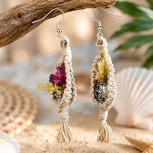 May include: A pair of macrame earrings featuring off-white knotted cord in a teardrop shape. Each earring holds a small silver-toned metal bead and dried flowers, with one displaying deep magenta petals and the other featuring yellow-toned dried flora.