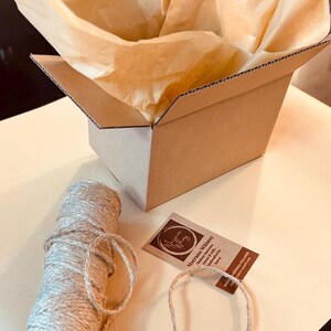 May include: A brown cardboard box with tissue paper, a spool of twine, and a tag. The box is open, revealing the tissue paper inside. The twine is beige and wound into a roll. The tag has text and a logo.