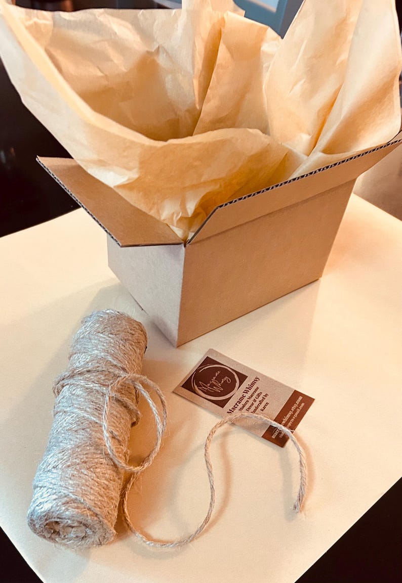 May include: A brown cardboard box with tissue paper, a spool of twine, and a tag. The box is open, revealing the tissue paper inside. The twine is beige and wound into a roll. The tag has text and a logo.