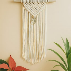 May include: Cream-colored macrame wall hanging with a wooden dowel. The design features a diamond-shaped knot pattern and a decorative stone pendant. The wall decor is displayed against a neutral background, with plants in the foreground.