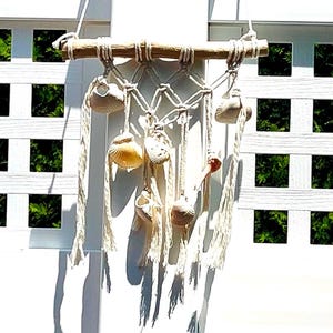 May include: A macrame wall hanging featuring a natural wooden dowel, cream-colored rope, and various seashells. The shells are in shades of beige, tan, and cream. The design includes knotted rope and hanging strands, creating a coastal, beachy aesthetic.