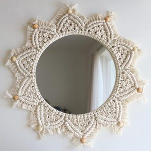 May include: A round mirror with a cream-colored macrame sunburst design. The mirror's frame features intricate knotting and fringe details, with wooden beads evenly spaced around the perimeter. The mirror is mounted on a white wall.