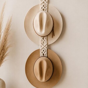 May include: A macrame hat rack with two hats. The hats are beige and brown, with a leather band. The rack is made of woven cream-colored rope and has a wooden ring at the top for hanging.