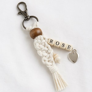 Op de afbeelding: Een crèmekleurige macramé sleutelhanger met de naam "ROSE" in houten letterkralen. Het heeft een houten kraal, een zilveren hartbedel en een metalen sluiting.