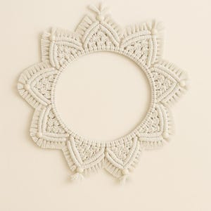 May include: Off-white macrame wall hanging in a sunburst design. The circular frame features intricate knotting patterns, beaded accents, and fringe detailing. This handcrafted piece adds a bohemian touch to home decor.