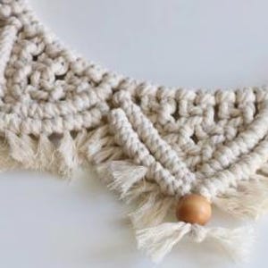 May include: Close-up of a cream-colored macrame wall hanging with a geometric design. The macrame features knotted patterns, fringe, and wooden beads. The piece is displayed against a white background, highlighting the texture and detail of the handcrafted item.