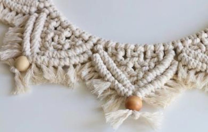 May include: Close-up of a cream-colored macrame wall hanging with a geometric design. The macrame features knotted patterns, fringe, and wooden beads. The piece is displayed against a white background, highlighting the texture and detail of the handcrafted item.