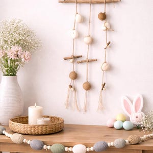 May include: An Easter-themed wall decoration featuring hanging spheres in white and brown, suspended from twine. A wooden table displays a candle, a woven tray, and a garland of pastel-coloured eggs. A bunny decoration and flowers complete the scene.
