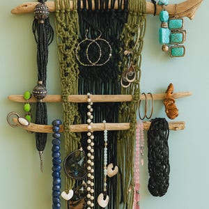 May include: A macrame jewelry organizer with a driftwood base, holding various necklaces, earrings, and bracelets. The macrame is black and olive green. Jewelry includes turquoise, pearl, and black beaded necklaces, and gold hoop earrings.