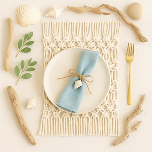 May include: A coastal-themed table setting with a white plate, light blue napkin tied with twine and a seashell, and a gold fork. The setting includes a macrame placemat, seashells, driftwood, and greenery, creating a beachy aesthetic.