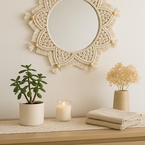 May include: A round macrame mirror with a cream-colored woven design and wooden bead accents hangs above a wooden dresser. A potted plant, a lit candle, and folded beige linens are on the dresser.