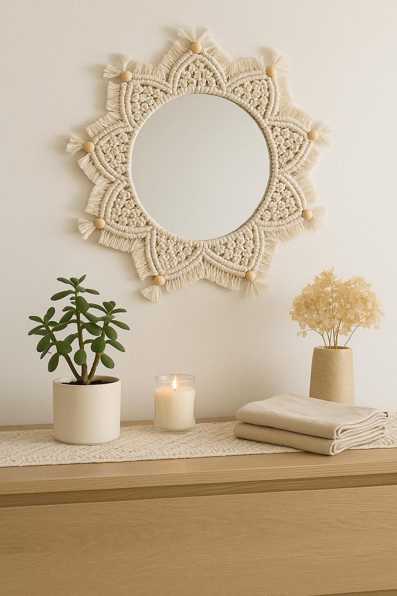 May include: A round macrame mirror with a cream-colored woven design and wooden bead accents hangs above a wooden dresser. A potted plant, a lit candle, and folded beige linens are on the dresser.