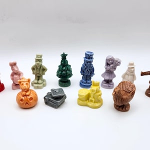 Puede incluir: Una colección de pequeñas figuras moldeadas de varios colores y formas, incluyendo una figura roja, un conejo rosa, un árbol verde, una figura azul, un castillo de arena amarillo, un pavo marrón, un espantapájaros marrón y libros grises.
