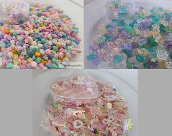 May include: Assortment of colourful resin flower embellishments in various pastel colours, including pink, blue, green, and white. The embellishments are in a clear container and on a white surface.  *decofrogcrafts