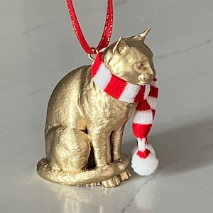 May include: A gold-colored cat ornament with a red and white striped scarf and a red ribbon for hanging. The cat is in a seated position, and the scarf has a white pom-pom. The ornament is likely for the holiday season.