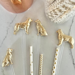 May include: Six gold dog-shaped cocktail stirrers with clear and patterned straws. The stirrers are on a white marble surface.