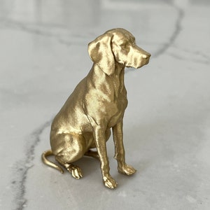 Puede incluir: Figura de perro dorada en posición sentada. El perro tiene un acabado metálico liso y está sentado erguido con la cabeza ligeramente girada hacia un lado. La figura está sobre una superficie clara.