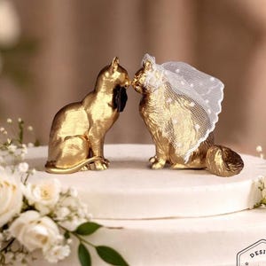 May include: A three-tiered white wedding cake decorated with white roses, greenery, and a pearl bead border. The top tier features two gold cat figurines, one wearing a veil. The cake has a "Design Nivera" logo.