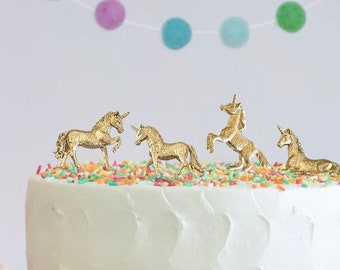 Peut inclure: Un gâteau blanc avec des décorations de gâteau en forme de licorne dorées et des vermicelles colorés sur le dessus. Le gâteau est sur un support à gâteau blanc.