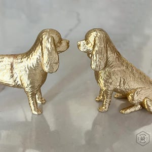 May include: Two gold-colored dog figurines, one standing and one sitting, face each other. The dogs have long, floppy ears and detailed fur texture. The figurines are on a light-colored surface.
