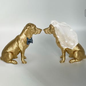 Pode incluir: Duas estatuetas de cães dourados, uma com uma gravata borboleta azul e a outra com um véu branco. Os cães estão virados um para o outro, sugerindo um tema de casamento. As estatuetas estão numa superfície branca.