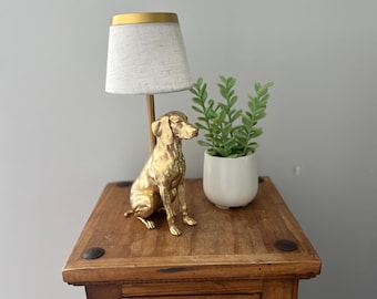Könnte beinhalten: Eine Tischlampe mit einem goldenen Hund als Lampenfuß und einem weißen Stoffschirm mit goldenem Rand. Daneben steht ein kleiner weißer Topf mit einer grünen Sukkulente. Das Logo Design Rivery ist auf dem Bild zu sehen.