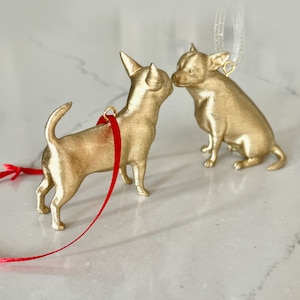 May include: Two gold-colored chihuahua ornaments, one with a red ribbon, are kissing. The ornaments are perfect for a Christmas tree or other holiday decor.