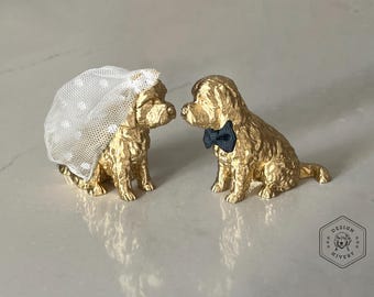 Könnte beinhalten: Zwei goldfarbene Hundefiguren, eine mit weißem Schleier mit Punkten und die andere mit einer marineblauen Fliege. Die Hunde blicken sich an, was auf ein Hochzeitsthema hindeutet. Die Figuren stehen auf einer hellen Oberfläche.