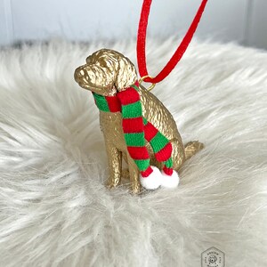 K&ouml;nnte beinhalten: Goldfarbenes Hundefiguren-Ornament mit einem rot-gr&uuml;nen gestreiften Schal. Das Ornament hat eine goldene Schlaufe zum Aufh&auml;ngen und wei&szlig;e Bommeln an den Enden des Schals.