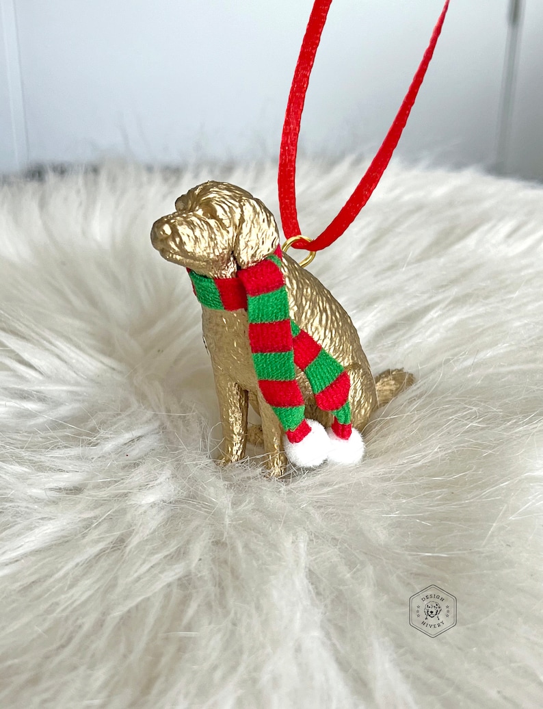 K&ouml;nnte beinhalten: Goldfarbenes Hundefiguren-Ornament mit einem rot-gr&uuml;nen gestreiften Schal. Das Ornament hat eine goldene Schlaufe zum Aufh&auml;ngen und wei&szlig;e Bommeln an den Enden des Schals.