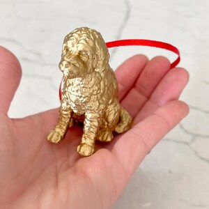 K&ouml;nnte beinhalten: Ein goldfarbener Hundefiguren-Anh&auml;nger, der mit gekreuzten Beinen sitzt und ein rotes Band um den Hals hat.