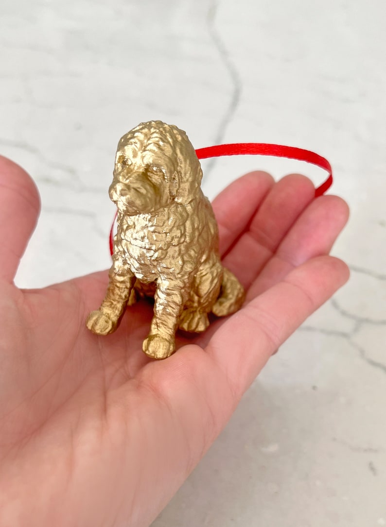 K&ouml;nnte beinhalten: Ein goldfarbener Hundefiguren-Anh&auml;nger, der mit gekreuzten Beinen sitzt und ein rotes Band um den Hals hat.