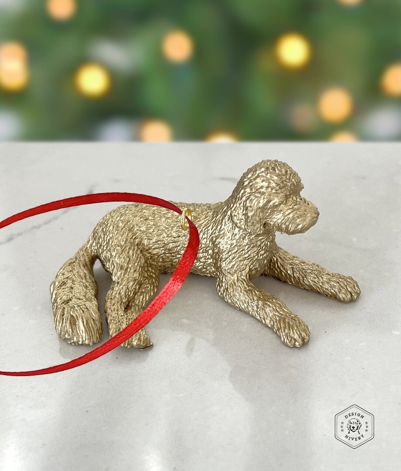 K&ouml;nnte beinhalten: Goldfarbenes Hundefiguren-Ornament mit rotem Band. Das Ornament hat die Form eines liegenden Hundes.