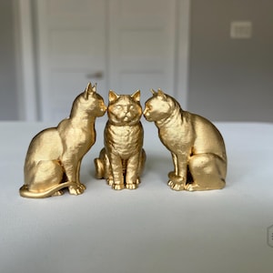 May include: Three gold-colored cat figurines. Two cats are seated, facing each other, while the third cat sits upright in the center. The cats have a textured surface and are positioned on a white surface.