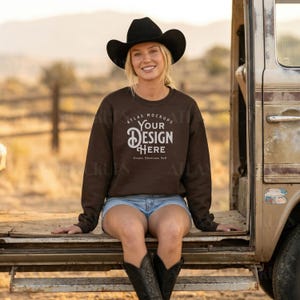 May include: A woman wearing a black cowboy hat, brown sweatshirt, and denim shorts. The sweatshirt has the text "YOUR DESIGN HERE" in white. She is sitting on the back of a vintage vehicle.