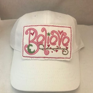 May include: White baseball cap with a pink embroidered patch that says "Believe" with a green leaf and pink hearts.