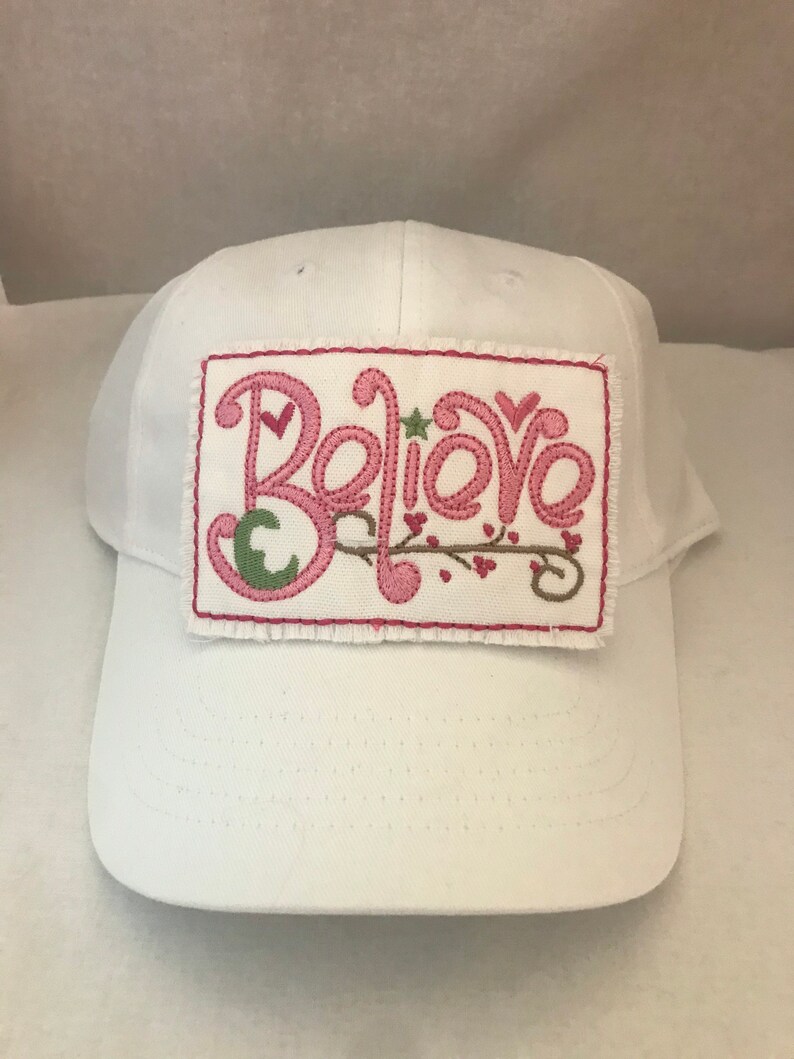 May include: White baseball cap with a pink embroidered patch that says "Believe" with a green leaf and pink hearts.