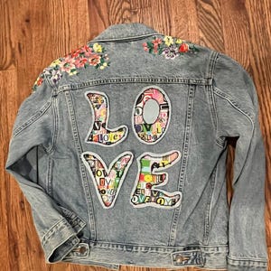 May include: A light blue denim jacket with a large "LOVE" patch on the back. The patch is made of colorful fabric scraps and is surrounded by embroidered flowers.