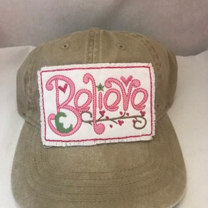 May include: A tan canvas baseball cap with a white patch that says "Believe" in pink and green embroidery.