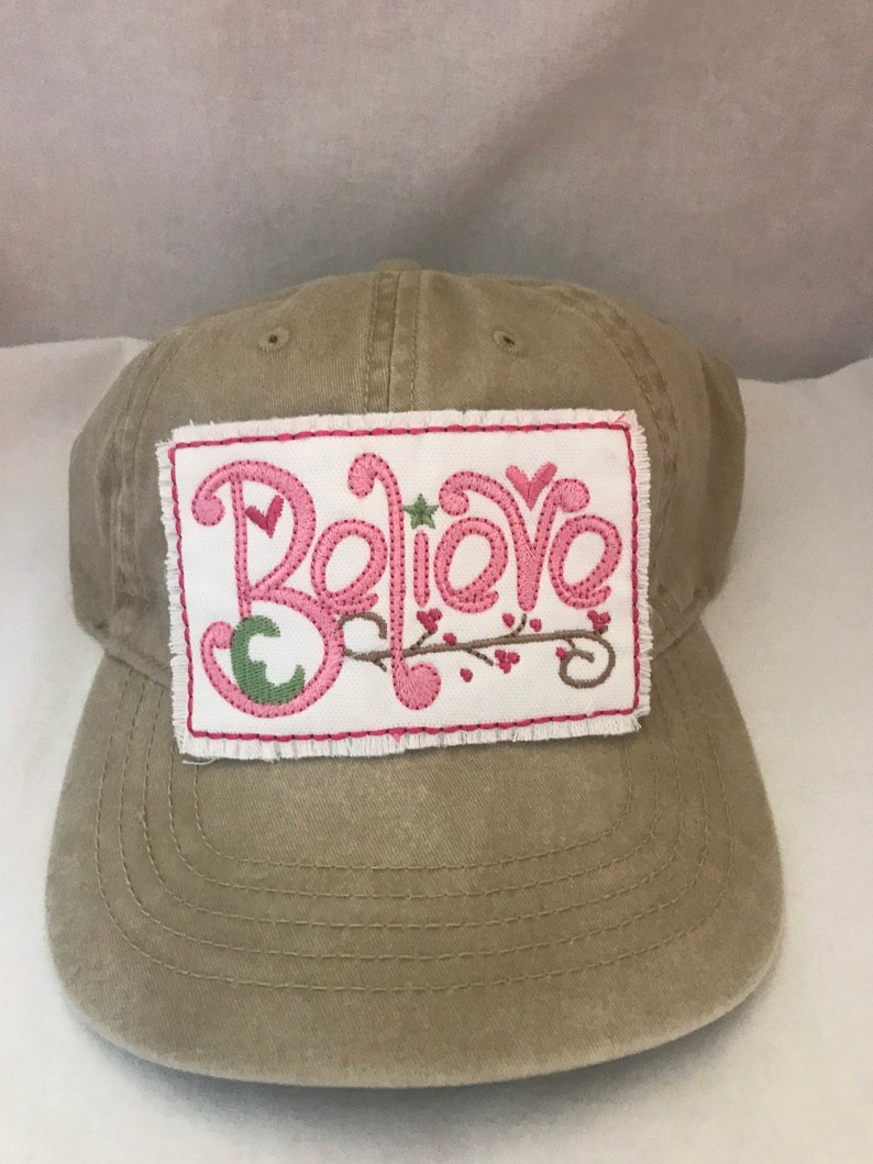 May include: A tan canvas baseball cap with a white patch that says "Believe" in pink and green embroidery.