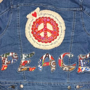 May include: A blue denim jacket featuring a large circular patch on the back with a red and orange peace sign, a small red heart, and the embroidered text All you need is love. Below, the word PEACE is appliqued in multi-colored patterned fabric.