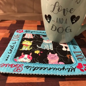 May include: A blue ceramic mug with the text "All you need is Love... and a DOG" in black lettering. The mug has two paw print hearts on the front. The mug is sitting on a black and white fabric coaster with a blue border and the text "All you need is... and a DOG" in white lettering. The coaster features a variety of colorful dog illustrations.