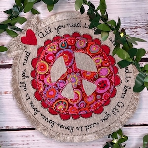 May include: A round, beige fabric patch with a red peace sign in the center. The peace sign is surrounded by a red scalloped border. The fabric patch has frayed edges and the text "All you need is love is love is all you need" is embroidered around the peace sign.