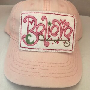 May include: Pink baseball cap with a white patch that says "Believe" in pink embroidered letters with green leaves and pink hearts.