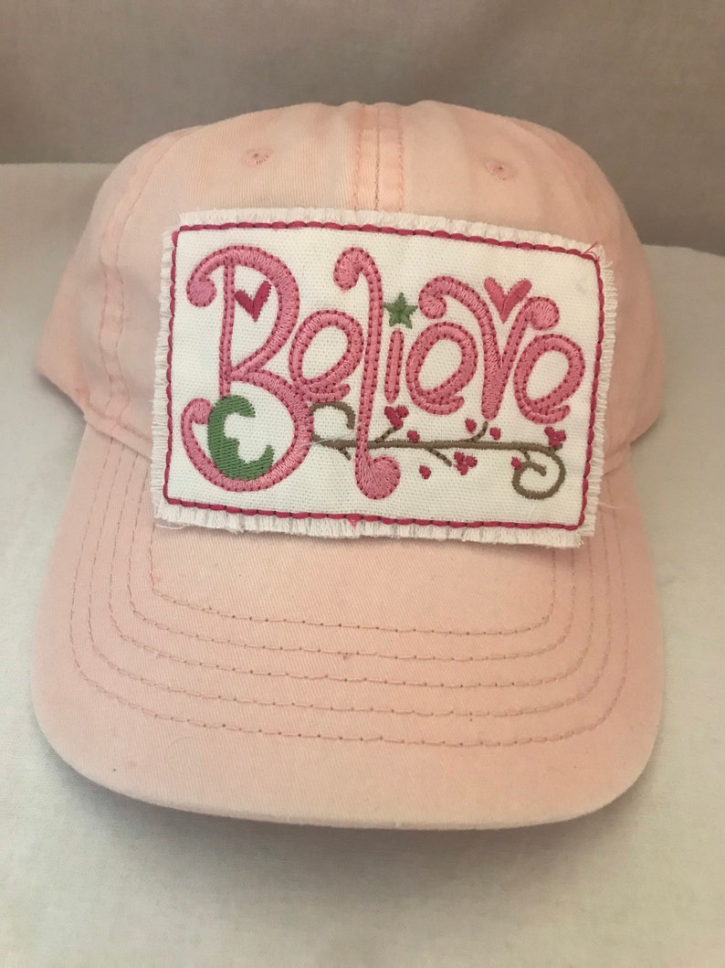 May include: Pink baseball cap with a white patch that says "Believe" in pink embroidered letters with green leaves and pink hearts.