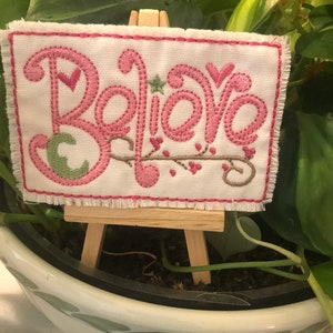 May include: A small white canvas with pink embroidery that reads "Believe" is displayed on a miniature wooden easel. The canvas is sitting on top of a potted plant.
