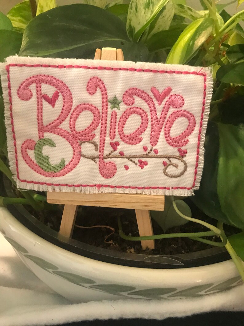 May include: A small white canvas with pink embroidery that reads "Believe" is displayed on a miniature wooden easel. The canvas is sitting on top of a potted plant.