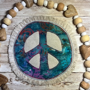 May include: A round, fringed peace sign with a teal and purple tie-dye pattern, stitched with red thread. The peace sign is surrounded by a beaded garland with wooden beads and heart-shaped accents.