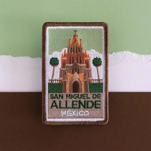 May include: Embroidered patch featuring the San Miguel de Allende church in Mexico. The patch has a brown border and depicts the church in shades of orange, brown, and green. The text "SAN MIGUEL DE ALLENDE MEXICO" is displayed.